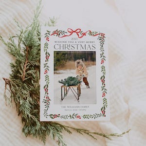 Pode incluir: Um cartão de Natal com uma foto de uma criança com roupa de inverno. O cartão tem uma borda decorativa de folhas verdes e bagas vermelhas. O texto diz "Wishing You a Very Merry Christmas" e "The Williams Family".
