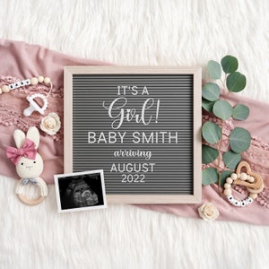 May include: A gray letter board with the text "It's a Girl! Baby Smith arriving August 2022". The board is surrounded by pink fabric, eucalyptus leaves, white flowers, and a baby rattle.