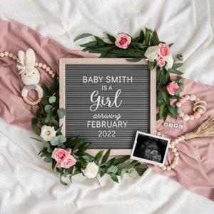 May include: A gray letter board with the text "Baby Smith is a Girl arriving February 2022". The board is surrounded by greenery, pink flowers, and a wooden bead garland. There is also a black and white ultrasound image and a wooden toy.