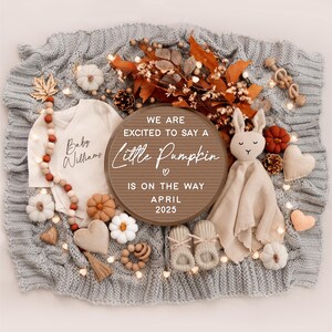 May include: A wooden sign with the text "We are excited to say a little pumpkin is on the way April 2025" surrounded by autumn-themed baby items, including a onesie, booties, a stuffed bunny, and a garland of wooden beads.