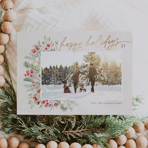 May include: A Christmas card with a watercolor border of greenery and red berries. The card features a photo of a family of three walking in the snow. The text "happy holidays 21" is written in gold script. The card is signed "love, the williams family".