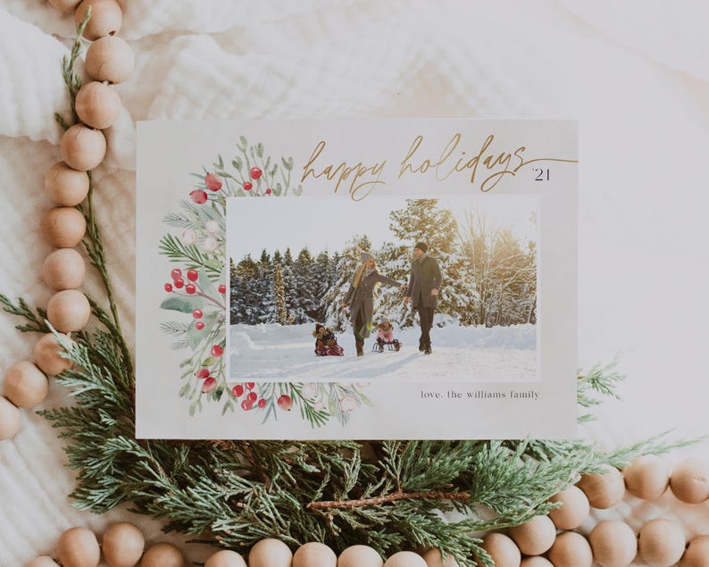 May include: A Christmas card with a watercolor border of greenery and red berries. The card features a photo of a family of three walking in the snow. The text "happy holidays 21" is written in gold script. The card is signed "love, the williams family".