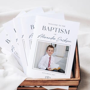 May include: White and black program for a baptism ceremony. The program features a photo of a young boy in a white shirt and red tie, holding a book. The text reads "Welcome to the Baptism of Alexander Jackson, September 9, 2023".