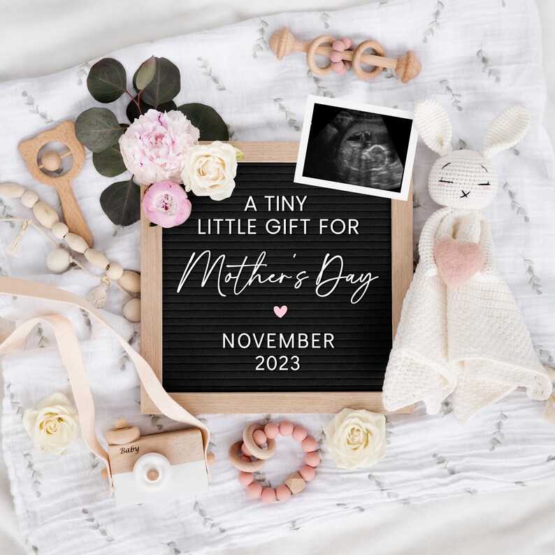 May include: A black letter board with the text "A tiny little gift for Mother's Day November 2023". A pink heart is above the date. The board is surrounded by a white blanket, a pink and white crocheted bunny, a wooden toy camera, a wooden rattle, a wooden ring, and a pink and white flower arrangement.
