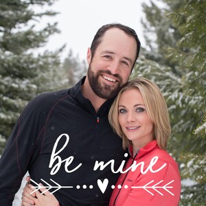 May include: A couple is smiling and hugging in front of a snowy forest. The man is wearing a black shirt and the woman is wearing a pink shirt. The text "be mine" is written in white cursive with a heart and arrows.