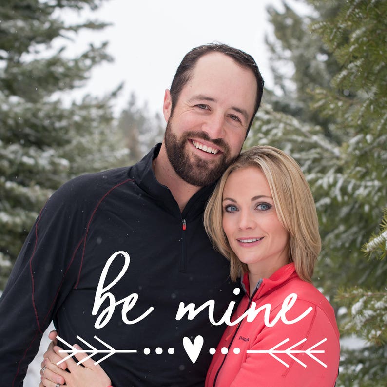 May include: A couple is smiling and hugging in front of a snowy forest. The man is wearing a black shirt and the woman is wearing a pink shirt. The text "be mine" is written in white cursive with a heart and arrows.