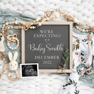 Puede incluir: Un tablero de letras gris con el texto "We're Expecting! Baby Smith December 2022". El tablero está rodeado de una manta blanca, una manta verde, cuentas de madera, un juguete de madera, un conejito de crochet y una foto de ecografía.