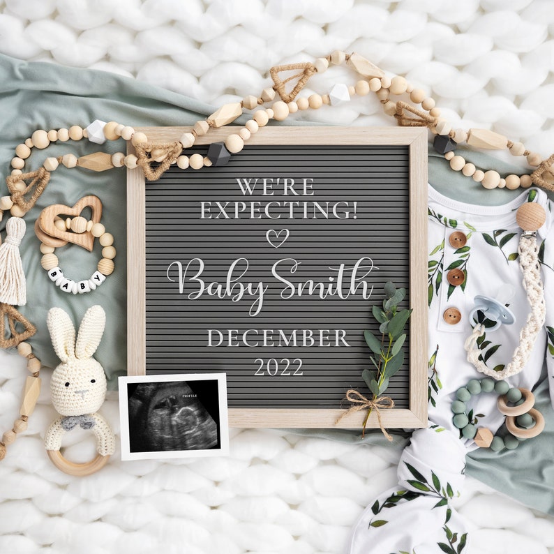 May include: A gray letter board with the text "We're Expecting! Baby Smith December 2022". The board is surrounded by a white blanket, a green blanket, wooden beads, a wooden toy, a crocheted bunny, and a sonogram photo.