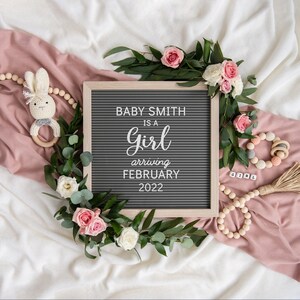 May include: A gray letter board with the text "Baby Smith is a girl arriving February 2022". The board is surrounded by greenery, pink roses, and a wooden bead garland.