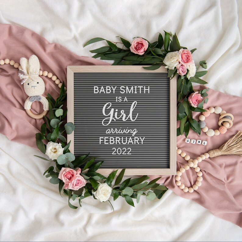 May include: A gray letter board with the text "Baby Smith is a girl arriving February 2022". The board is surrounded by greenery, pink roses, and a wooden bead garland.