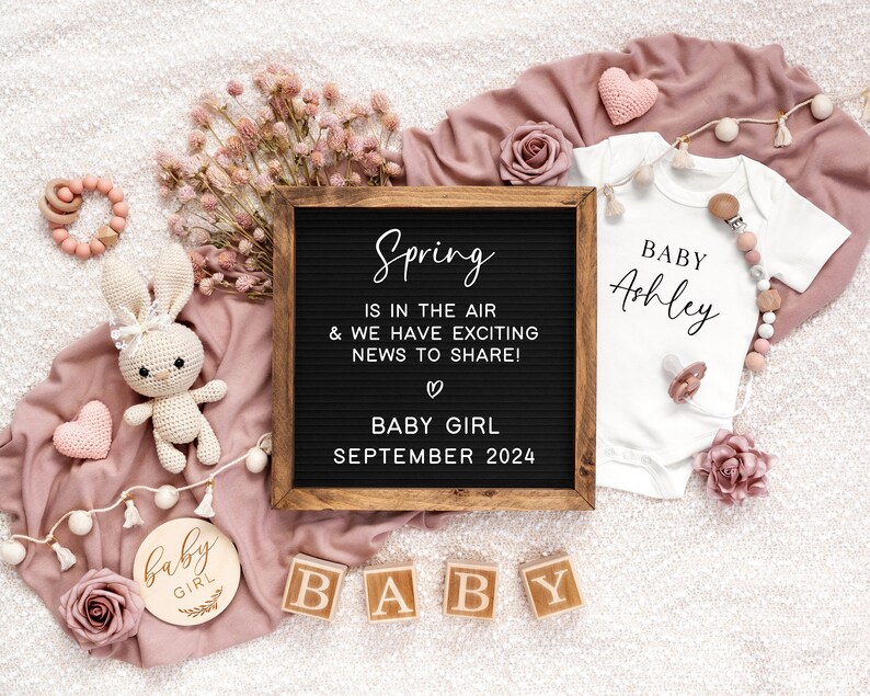May include: A pink and white baby announcement with a chalkboard sign that reads "Spring is in the air & we have exciting news to share! Baby Girl September 2024". There is a white onesie with the text "Baby Ashley" and a wooden sign that reads "baby girl".