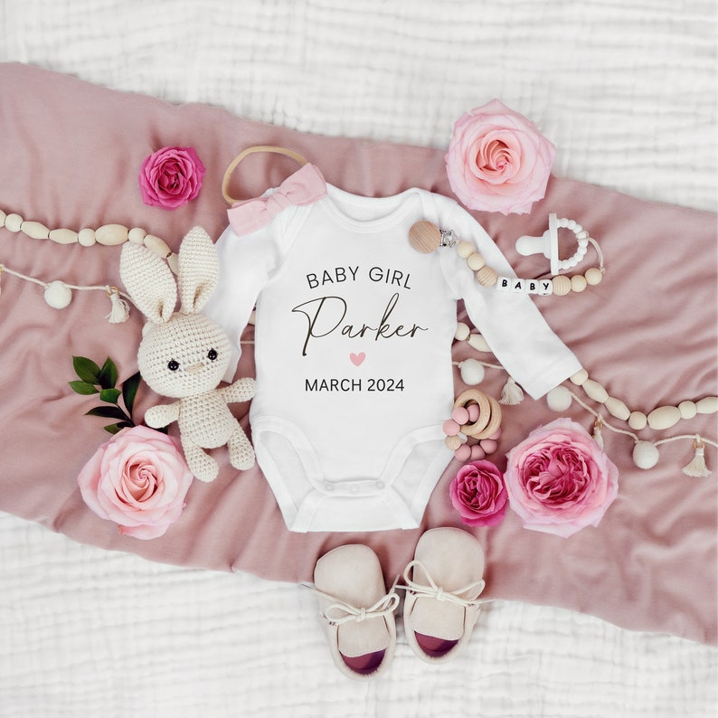 May include: White baby onesie with the text "Baby Girl Parker" and a pink heart, with the date "March 2024". The onesie is surrounded by pink roses, a crocheted bunny, wooden beads, and baby shoes.