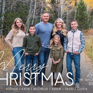May include: A family of seven poses for a Christmas photo in a forest setting. The family is dressed in casual attire and smiles for the camera. The text "Merry Christmas" and "Love, The Taylor Family - 2021 - Joshua | Katie | Michelle | Adam | Dean | Olivia" is written below the family.