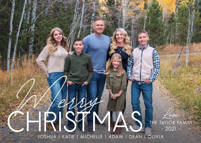 May include: A family of seven poses for a Christmas photo in a forest setting. The family is dressed in casual attire and smiles for the camera. The text "Merry Christmas" and "Love, The Taylor Family - 2021 - Joshua | Katie | Michelle | Adam | Dean | Olivia" is written below the family.