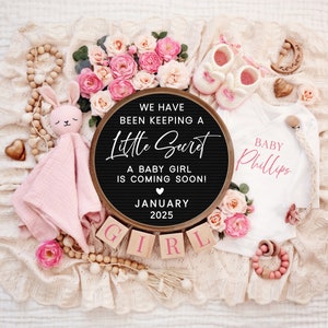 K&ouml;nnte beinhalten: Ein schwarzes Buchstabenbrett mit dem Text "We have been keeping a little secret. A baby girl is coming soon! January 2025."  Rosa und wei&szlig;e Babykleidung, eine rosa Hasenrassel und Holzbl&ouml;cke, die "GIRL" buchstabieren, sind um das Buchstabenbrett herum angeordnet.