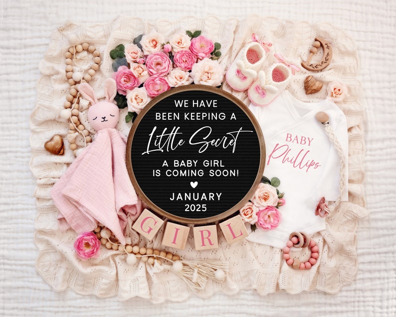 K&ouml;nnte beinhalten: Ein schwarzes Buchstabenbrett mit dem Text "We have been keeping a little secret. A baby girl is coming soon! January 2025."  Rosa und wei&szlig;e Babykleidung, eine rosa Hasenrassel und Holzbl&ouml;cke, die "GIRL" buchstabieren, sind um das Buchstabenbrett herum angeordnet.
