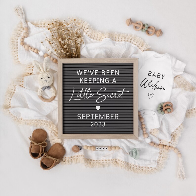 May include: A black letter board with the text "We've Been Keeping A Little Secret" and the date "September 2023". A white onesie with the text "Baby Wilson" is next to the letter board. There are also a few baby toys and a blanket in the image.