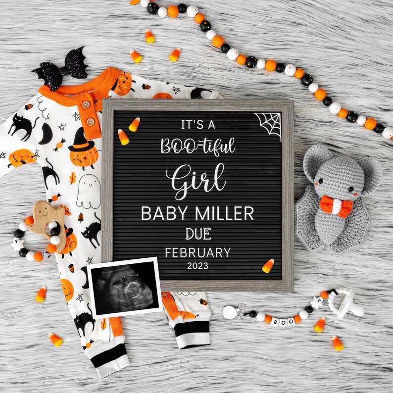 Puede incluir: Un tablero de letras negro con el texto "It's a BOO-tiful Girl Baby Miller Due February 2023".  Un murci&eacute;lago de crochet gris con una pajarita naranja est&aacute; en el lado derecho del tablero.  Un mameluco de beb&eacute; blanco y naranja con un estampado de Halloween negro y naranja est&aacute; en el lado izquierdo del tablero.  Hay ma&iacute;z dulce naranja y blanco y un collar de cuentas blanco y negro con un sonajero de fantasma de madera en el tablero.