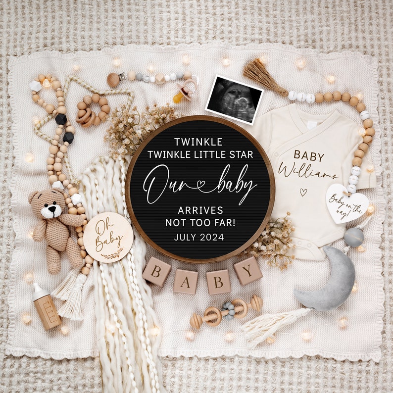 May include: A nursery announcement with a black letter board that reads "Twinkle Twinkle Little Star Our Baby Arrives Not Too Far! July 2024". There are wooden blocks that spell "Baby", a wooden sign that says "Oh Baby", a white onesie with the text "Baby Williams", a crocheted teddy bear, and a gray crescent moon.