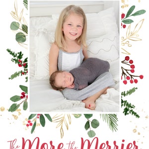 May include: A Christmas card featuring a watercolor illustration of greenery and red berries with gold accents. The card says "The More the Merrier" in red text and "THE MORGAN FAMILY THOMAS | KELLY | OLIVIA | GRAYSON - 2020 -" in black text. A photo of a young girl holding a baby is in the center of the card.