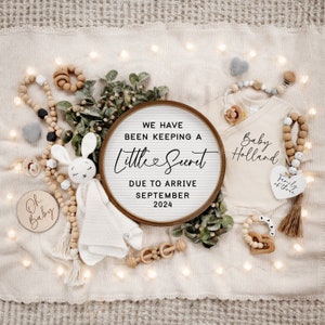 May include: A wooden sign with the text "We have been keeping a little secret due to arrive September 2024". The sign is surrounded by a white blanket, a baby onesie, a wooden toy, and a stuffed bunny.