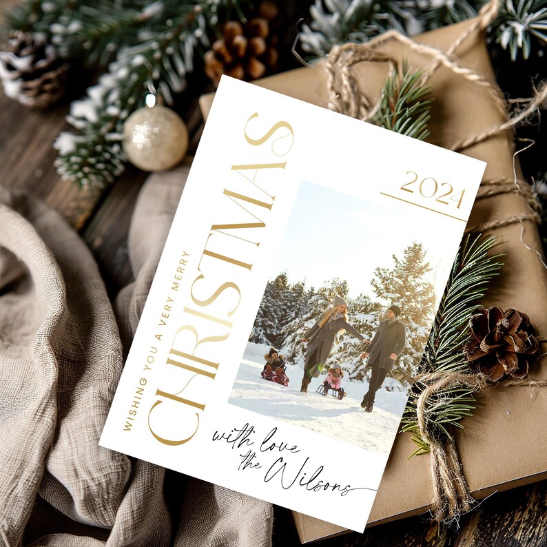 May include: A white Christmas card with gold lettering that reads "Wishing you a very merry Christmas 2021" and "with love the Wilsons". The card features a photo of a family of four in a snowy winter scene.