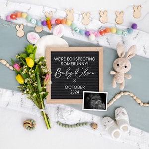 K&ouml;nnte beinhalten: Ein schwarzes Buchstabenbrett mit dem Text "We're eggspecting somebunny! Baby Olsen October 2024" umgeben von einer wei&szlig;en Decke, einem Strau&szlig; gelber und rosa Blumen, einem geh&auml;kelten Hasen, einer Hasen-Girlande aus Holz, einer Perlenkette aus Holz, einer Babysocke, einem Schnuller und einem Ultraschallbild.