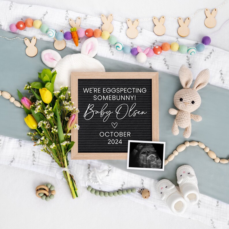 K&ouml;nnte beinhalten: Ein schwarzes Buchstabenbrett mit dem Text "We're eggspecting somebunny! Baby Olsen October 2024" umgeben von einer wei&szlig;en Decke, einem Strau&szlig; gelber und rosa Blumen, einem geh&auml;kelten Hasen, einer Hasen-Girlande aus Holz, einer Perlenkette aus Holz, einer Babysocke, einem Schnuller und einem Ultraschallbild.