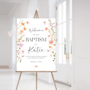 May include: A welcome sign for a baptism with a floral design in shades of pink and orange. The sign reads "Welcome to the Baptism of Katie The Church of Jesus Christ of Latter-day Saints March 1, 2025".