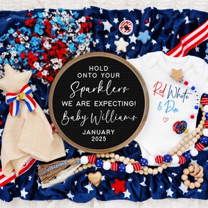 Op de afbeelding: Een zwart letterbord met de tekst "Hold onto your sparklers, we are expecting! Baby Williams, January 2025". Een wit rompertje met de tekst "Red, White & Due" en een rood hart. Een rood-wit-blauwe sterrendek met een stervormig knuffeldier.