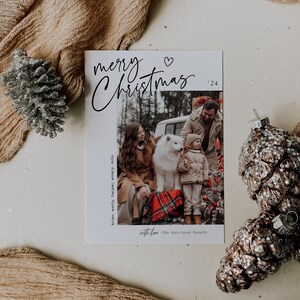May include: A white Christmas card with black calligraphy that reads "merry Christmas '24". A heart symbol is above the word "Christmas". A photo of a family with a dog is in the bottom half of the card. The text "brian, emily, harper, emery, milo" is on the left side of the photo. The text "with love the harrison family" is below the photo.