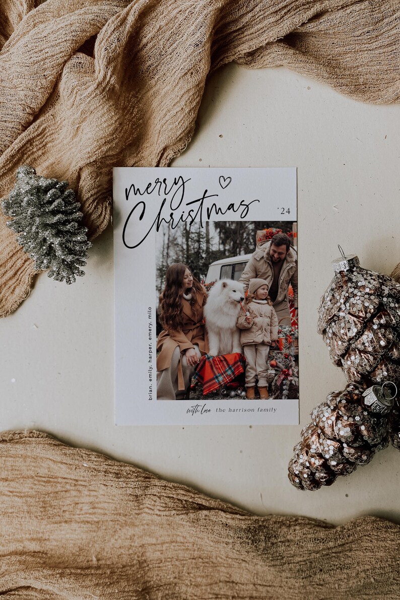 May include: A white Christmas card with black calligraphy that reads "merry Christmas '24". A heart symbol is above the word "Christmas". A photo of a family with a dog is in the bottom half of the card. The text "brian, emily, harper, emery, milo" is on the left side of the photo. The text "with love the harrison family" is below the photo.