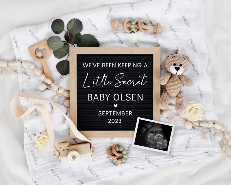 K&ouml;nnte beinhalten: Ein schwarzes Buchstabenbrett mit dem Text "We've been keeping a little secret Baby Olsen September 2023" umgeben von einer wei&szlig;en Decke, einem Teddyb&auml;r, Holzspielzeug und wei&szlig;en Rosen.