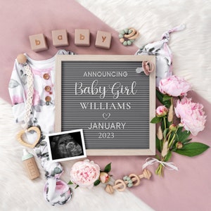 Puede incluir: Un pelele de beb&eacute; rosa y blanco con estampado floral, un tablero de letras que anuncia "Announcing Baby Girl Williams January 2023", un coraz&oacute;n de madera, un biber&oacute;n, un sonajero de madera, un chupete, un coraz&oacute;n de madera, una imagen de ecograf&iacute;a, un ramo de peon&iacute;as rosas y bloques de madera que deletrean "baby" sobre una superficie rosa y blanca.