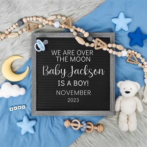 May include: A black letter board with the text "We are over the moon Baby Jackson is a boy! November 2023". The board is surrounded by blue fabric, a wooden bead garland, a yellow crescent moon, a white cloud, a blue star, a white teddy bear, and a wooden rattle.