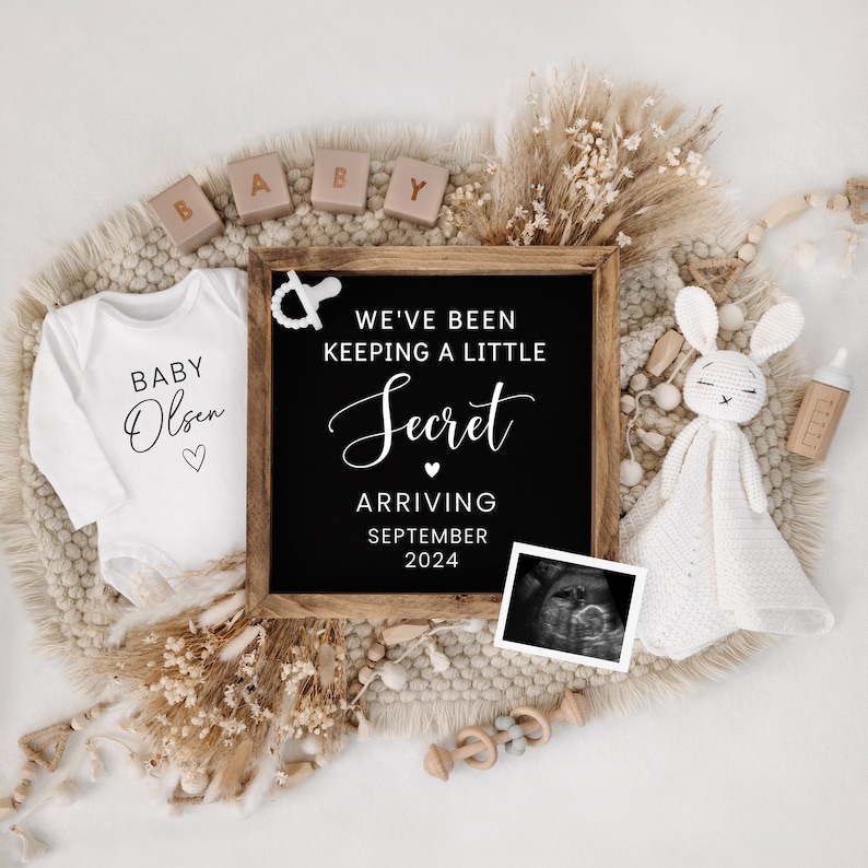 May include: A chalkboard sign with the text "We've been keeping a little secret. Arriving September 2024."  A white onesie with the text "Baby Olsen" is next to the sign.  There are wooden blocks that spell out "BABY" and a crocheted bunny with a white blanket.
