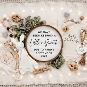 May include: A wooden sign with the text "We have been keeping a little secret due to arrive September 2024". The sign is surrounded by a variety of baby items, including a knitted bunny, a wooden rattle, and a white onesie with the text "Baby Richards" and "Family of three".