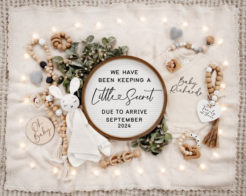 May include: A wooden sign with the text "We have been keeping a little secret due to arrive September 2024". The sign is surrounded by a variety of baby items, including a knitted bunny, a wooden rattle, and a white onesie with the text "Baby Richards" and "Family of three".