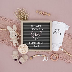 Op de afbeelding: Een roze en witte babymeisjesaankondiging met een letterbord waarop staat "We are having a girl September 2023". Er is een wit rompertje met de tekst "Baby Olivia" en andere babyartikelen zoals een houten rammelaar, een speen en een paar babyschoentjes.