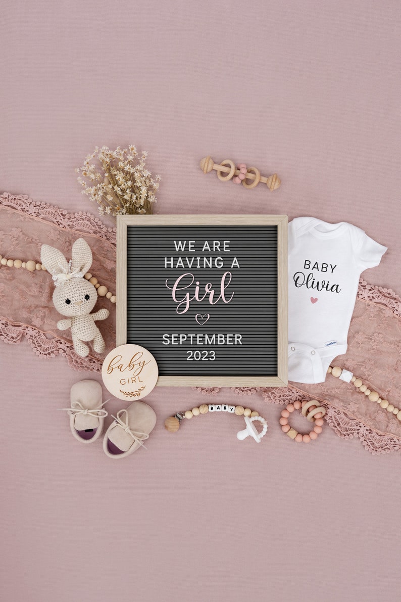 Op de afbeelding: Een roze en witte babymeisjesaankondiging met een letterbord waarop staat "We are having a girl September 2023". Er is een wit rompertje met de tekst "Baby Olivia" en andere babyartikelen zoals een houten rammelaar, een speen en een paar babyschoentjes.