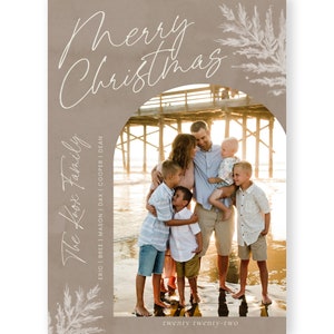 May include: A Christmas card featuring a family of five standing on a beach in front of a pier. The card has a brown background with white text that reads "Merry Christmas" and "The Koox Family" with the names of the family members listed below. The text "twenty twenty-two" is at the bottom of the card.