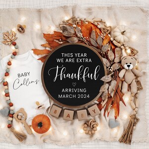 May include: A black letter board with the text "This year we are extra thankful arriving March 2024". The board is surrounded by fall-themed decorations, including a white onesie with the text "Baby Collins", a crocheted teddy bear, and a string of lights.