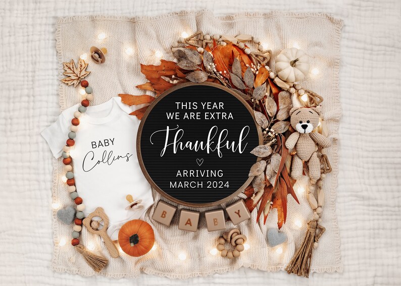 May include: A black letter board with the text "This year we are extra thankful arriving March 2024". The board is surrounded by fall-themed decorations, including a white onesie with the text "Baby Collins", a crocheted teddy bear, and a string of lights.