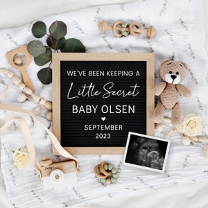 K&ouml;nnte beinhalten: Ein schwarzes Buchstabenbrett mit wei&szlig;em Text, der "We've been keeping a little secret, Baby Olsen, September 2023" ank&uuml;ndigt. Das Brett ist von Babyspielzeug, einem Teddyb&auml;r und einem Ultraschallbild umgeben.