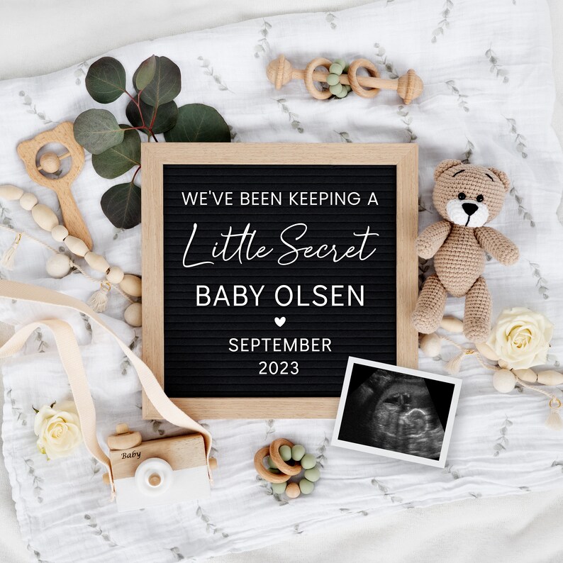 K&ouml;nnte beinhalten: Ein schwarzes Buchstabenbrett mit wei&szlig;em Text, der "We've been keeping a little secret, Baby Olsen, September 2023" ank&uuml;ndigt. Das Brett ist von Babyspielzeug, einem Teddyb&auml;r und einem Ultraschallbild umgeben.