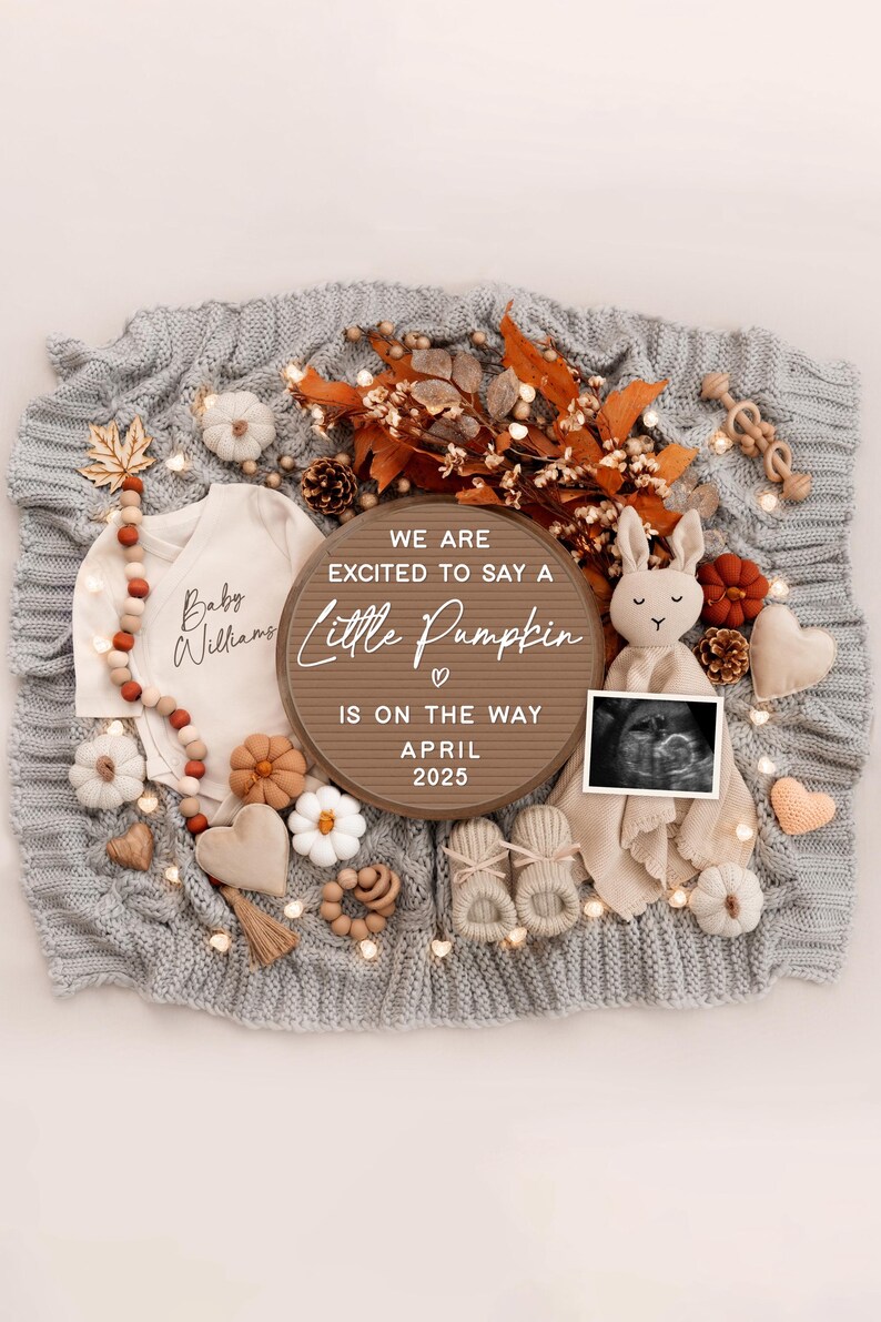 May include: A wooden sign with the text "We are excited to say a little pumpkin is on the way April 2025". The sign is surrounded by autumn-themed baby items, including a knitted blanket, a baby onesie, a stuffed bunny, and a pair of booties.