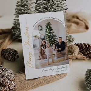 May include: A white Christmas card with a gold inscription that reads "Merry Christmas". The card features a photo of a family of three standing in front of a Christmas tree. The text "Wishing you a joyous holiday season!" is at the top of the card. The text "xoxo the jackson family twenty twenty-four" is at the bottom of the card.