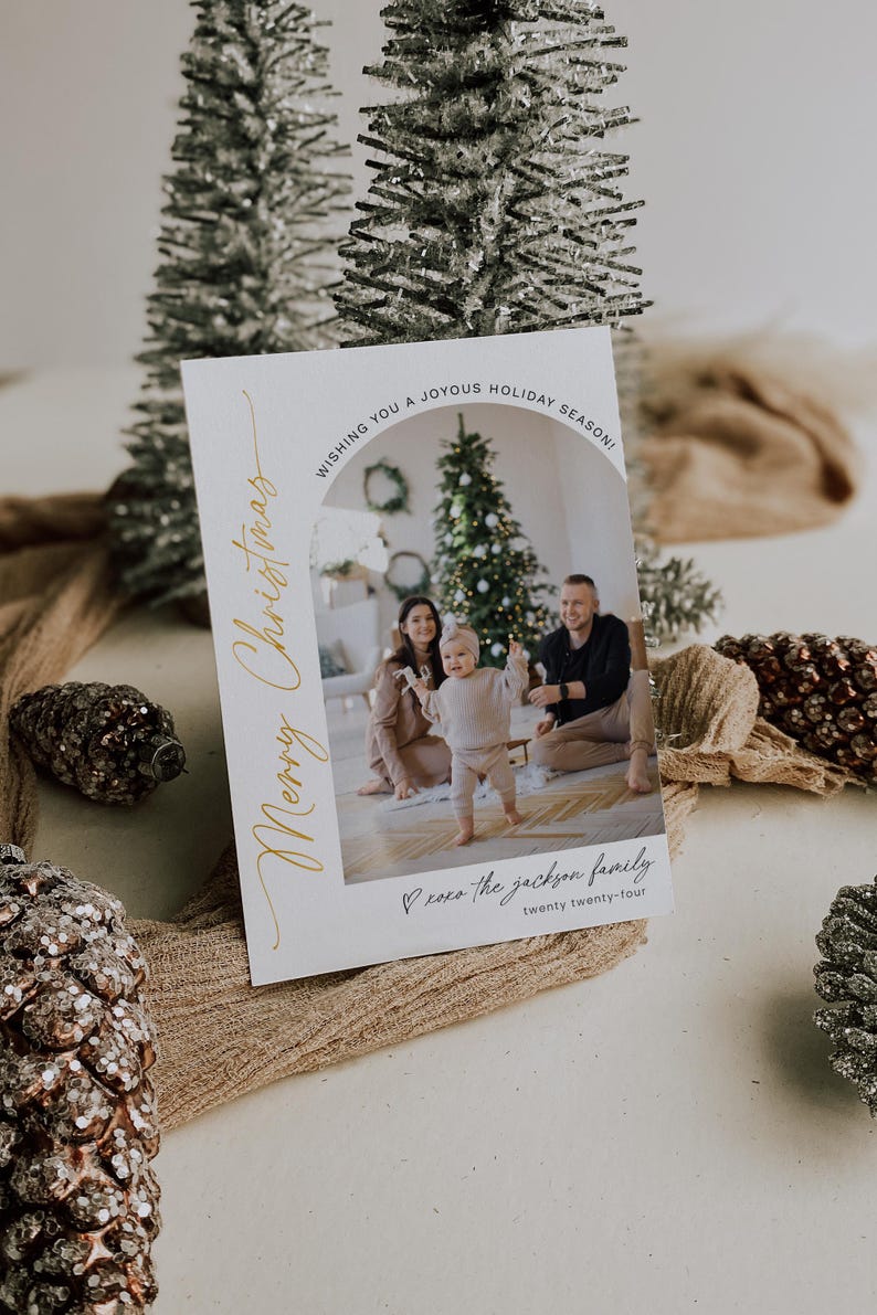 May include: A white Christmas card with a gold inscription that reads "Merry Christmas". The card features a photo of a family of three standing in front of a Christmas tree. The text "Wishing you a joyous holiday season!" is at the top of the card. The text "xoxo the jackson family twenty twenty-four" is at the bottom of the card.