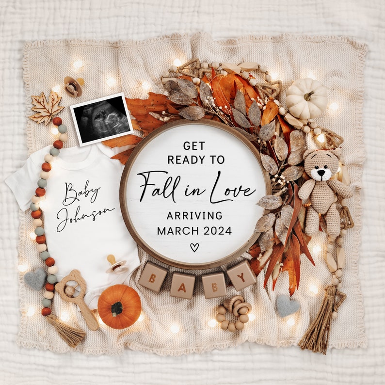 May include: A white babygrow with the text "Baby Johnson" on it, a wooden sign with the text "Get Ready to Fall in Love Arriving March 2024", and wooden blocks spelling out "BABY" on a tan blanket with autumn-themed decorations.