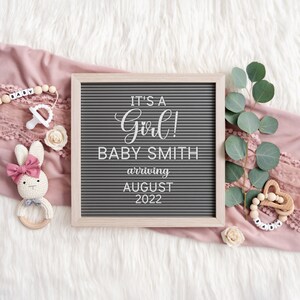 May include: A gray letter board with the text "It's a Girl! Baby Smith arriving August 2022". The board is surrounded by pink fabric, a wooden baby rattle, a white pacifier, and a crocheted bunny toy.